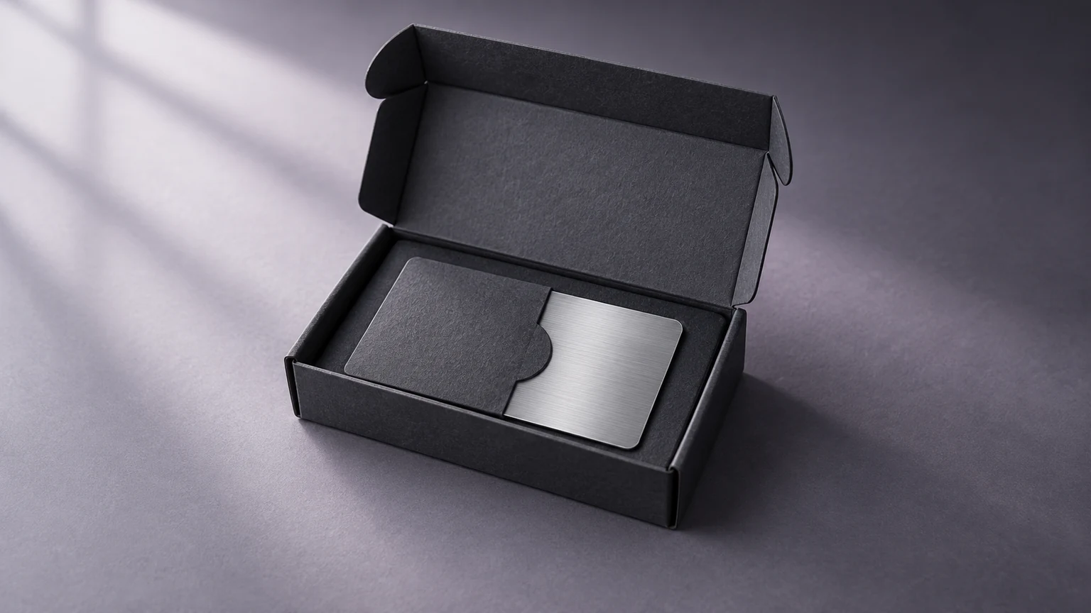 A brushed silver card in a protective sleeve inside an open graphite mailer, softly lit on a lavender-grey surface.